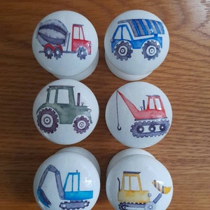 May include: Six white round knobs with different construction vehicle designs. The knobs feature a cement mixer, a dump truck, a tractor, a crane, an excavator, and a bulldozer.