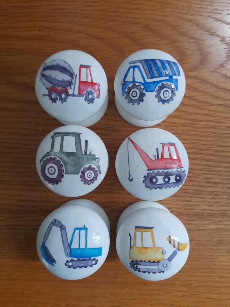 May include: Six white round knobs with different construction vehicle designs. The knobs feature a cement mixer, a dump truck, a tractor, a crane, an excavator, and a bulldozer.