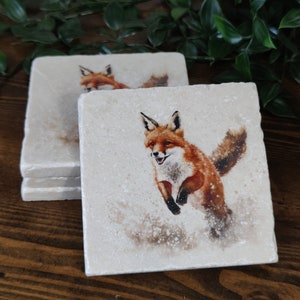 May include: Set of four square coasters featuring a watercolour illustration of a red fox jumping in the snow. The coasters are made of natural stone and have a white background.