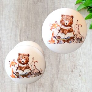 Pode incluir: Dois puxadores de armário de cerâmica branca com um design de animais da floresta. Cada puxador apresenta um urso castanho, uma raposa laranja e um coelho reunidos em torno de um livro aberto. Ideal para um quarto infantil.