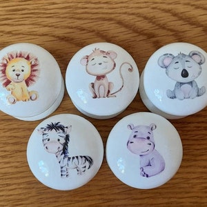 May include: Five white ceramic cabinet knobs with cute animal illustrations. The knobs feature a lion, monkey, koala, zebra, and hippo. Each knob has a round shape and is suitable for a nursery or children's room.
