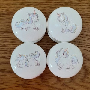 Może przedstawiać: Cztery białe, okrągłe drewniane pudełka z motywami jednorożców. Każde pudełko przedstawia jednorożca w innej pozie, w tym leżącego jednorożca, stojącego jednorożca i jednorożca z opuszczoną głową. Jednorożce są wszystkie białe z pastelowymi różowymi, niebieskimi i żółtymi akcentami.
