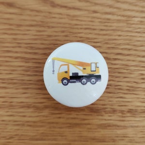 Op de afbeelding: Een witte knop met een illustratie van een gele kraanwagen. De truck heeft een zwart chassis en een gele giek.