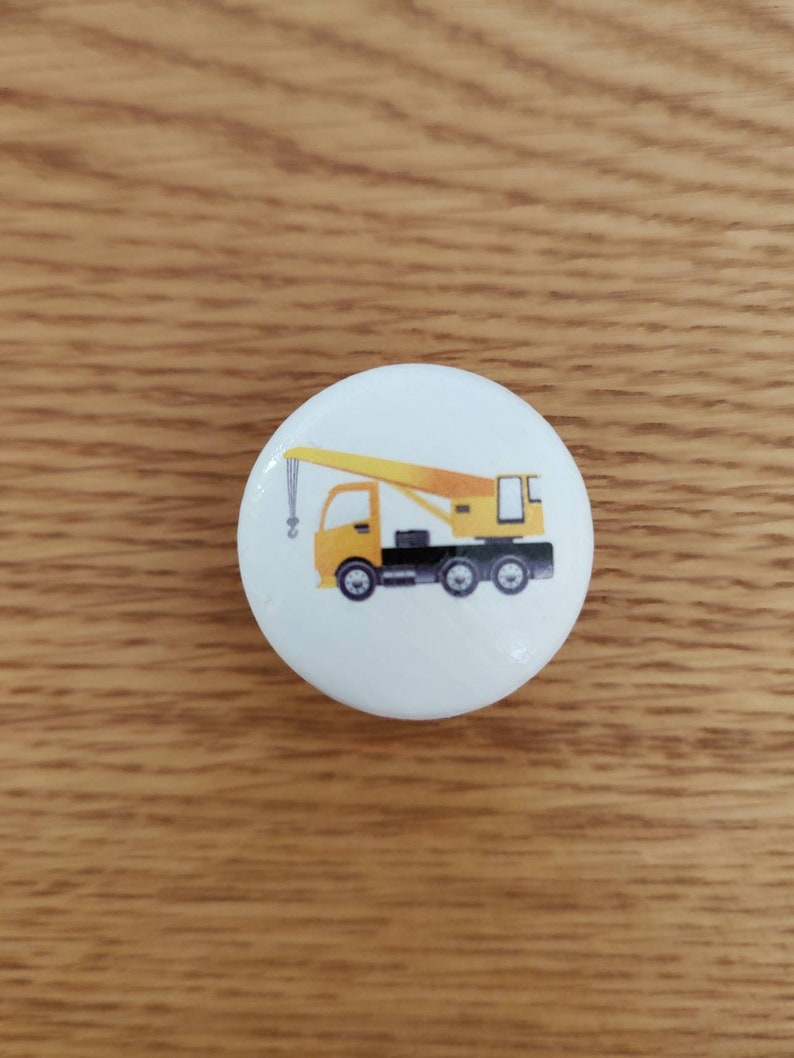 Op de afbeelding: Een witte knop met een illustratie van een gele kraanwagen. De truck heeft een zwart chassis en een gele giek.