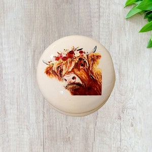 Peut inclure: Une boîte à bijoux ronde en céramique blanche avec une image peinte d'une vache écossaise portant une couronne de fleurs. La vache a une fourrure brune et un nez rose.