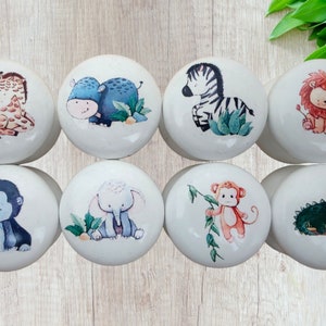 May include: Eight white ceramic cabinet knobs with colorful illustrations of safari animals, including a giraffe, hippo, zebra, lion, gorilla, elephant, monkey, and crocodile. Each knob features a different animal design.