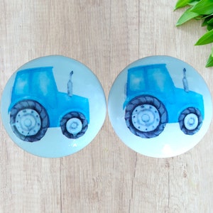 May include: Two round, light blue decorative objects with a watercolor-style image of a blue tractor. The tractor has dark blue tires and a light blue body. The background is a light blue color.