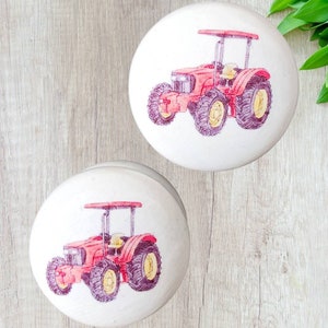 Op de afbeelding: Twee witte keramische knoppen met een rood tractorontwerp. De tractor is getekend in roze en geel.