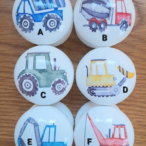 May include: Six white ceramic knobs with different construction vehicle designs. The knobs are labeled A through F. The vehicles include a blue dump truck, a red cement mixer, a green tractor, a yellow bulldozer, a blue excavator, and a red crane.