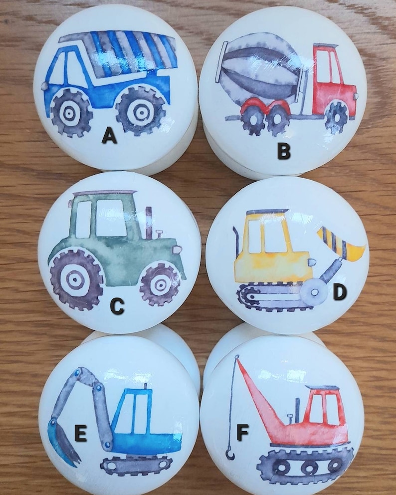 May include: Six white ceramic knobs with different construction vehicle designs. The knobs are labeled A through F. The vehicles include a blue dump truck, a red cement mixer, a green tractor, a yellow bulldozer, a blue excavator, and a red crane.