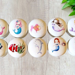 Könnte beinhalten: Zehn runde Holzgriffe mit bemalten Designs zum Thema Meer. Die Designs beinhalten Meerjungfrauen, Seepferdchen, Fische, Korallen und einen Delfin.