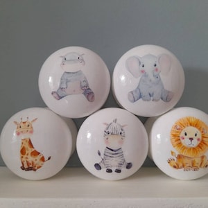 May include: Five white ceramic knobs with different animal designs. The knobs feature a giraffe, zebra, lion, elephant, and hippopotamus. Each knob has a watercolor-style design.