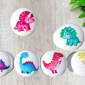 May include: Set of six white ceramic knobs with colourful dinosaur illustrations. The dinosaurs are a pink triceratops, a blue stegosaurus, a red T-Rex, a yellow dinosaur, a green triceratops, and a purple brachiosaurus.