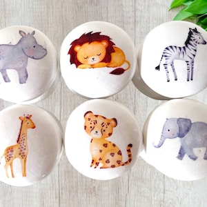 May include: Set of six white cabinet knobs featuring watercolor illustrations of safari animals: a rhino, lion, zebra, giraffe, cheetah, and elephant. Each knob has a unique animal design, perfect for a nursery or children's room.