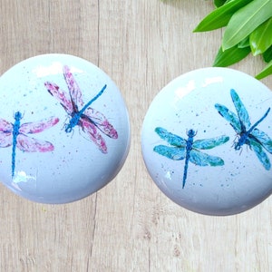 Peut inclure: Deux dessous-de-verre ronds en céramique blanche avec des motifs de libellules. Un dessous-de-verre présente deux libellules roses avec des ailes bleues, et l'autre dessous-de-verre présente deux libellules bleues avec des ailes vertes.