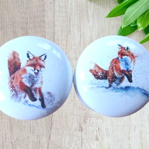 Puede incluir: Dos pomos de cerámica blanca con un diseño de zorro rojo. Los zorros se representan en un paisaje invernal nevado.