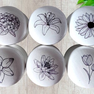 May include: Six white ceramic round containers with black floral designs. The designs include a hydrangea, a lily, a sunflower, a daffodil, a water lily, and a tulip.
