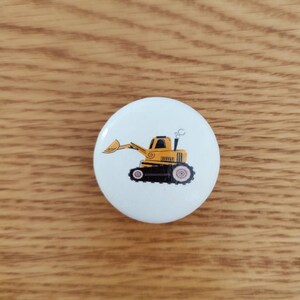 Op de afbeelding: Een witte ronde knop met een gele cartoon-graafmachine erop. De graafmachine heeft een zwarte cabine en zwarte wielen.