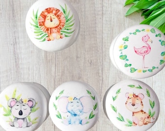 Puede incluir: Un conjunto de pomos de cerámica blanca con ilustraciones de acuarela de animales de la jungla. Los diseños incluyen un león, un flamenco, un koala, un elefante y un tigre, cada uno rodeado de vegetación.