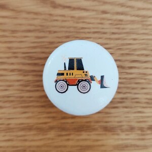 Op de afbeelding: Een witte keramische knop met een ontwerp van een gele en zwarte cartoon-graafmachine.