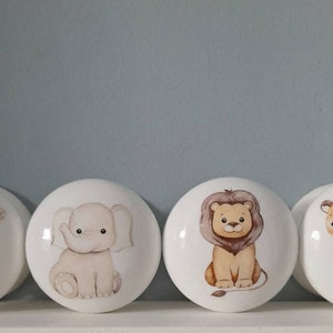 May include: Four white ceramic knobs with animal illustrations. The knobs feature a monkey, elephant, lion, and tiger. The animals are depicted in a cartoon style with brown, beige, and orange colors.