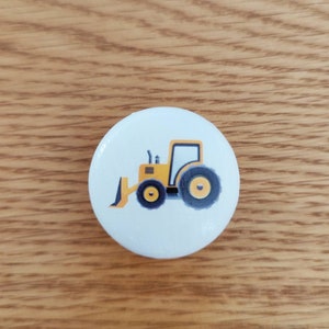 Op de afbeelding: Een witte keramische knop met een gele tractorillustratie. De tractor heeft zwarte wielen en een zwarte ploeg.