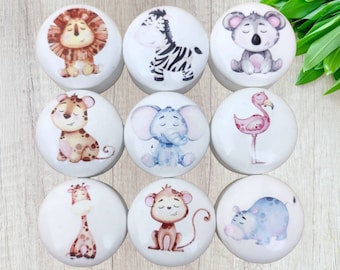 Puede incluir: Nueve pomos de cerámica blanca con diferentes ilustraciones de animales. Los animales incluyen un león, una cebra, un koala, un leopardo, un elefante, un flamenco, una jirafa, un mono y un hipopótamo. Todos los animales están representados en un estilo de dibujos animados con detalles como de acuarela.