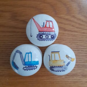 May include: Three ceramic knobs with a white background and colorful illustrations of construction vehicles. The knobs feature a red crane, a blue excavator, and a yellow bulldozer.