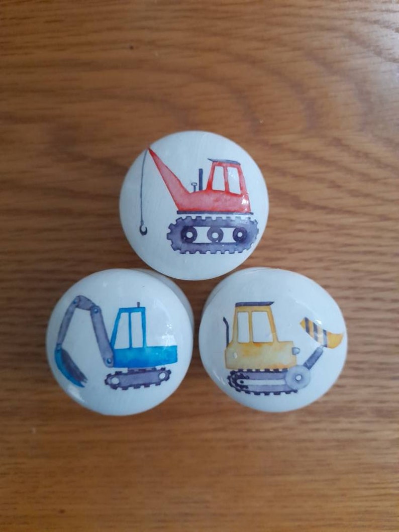 May include: Three ceramic knobs with a white background and colorful illustrations of construction vehicles. The knobs feature a red crane, a blue excavator, and a yellow bulldozer.