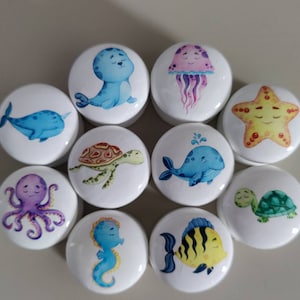 Könnte beinhalten: Eine Sammlung weißer Keramik-Schrankknöpfe mit bunten Cartoon-Meeresbewohnern. Designs umfassen einen Wal, eine Robbe, eine Qualle, einen Seestern, eine Schildkröte, einen Oktopus, ein Seepferdchen, einen Fisch und eine zweite Schildkröte und einen Wal. Jeder Knopf hat ein lächelndes Meereswesen.