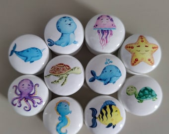 May include: A collection of white ceramic cabinet knobs featuring colorful, cartoon sea creatures. Designs include a whale, seal, jellyfish, starfish, turtle, octopus, seahorse, fish, and a second turtle and whale. Each knob has a unique, smiling sea creature.