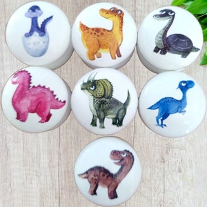 Może przedstawiać: Zestaw siedmiu białych ceramicznych gałek do szafek, z których każda przedstawia ilustrację dinozaura w akwareli. Dinozaury obejmują różowego stegozaura, zielonego triceratopsa i brązowego T-Rexa. Idealne do pokoju dziecka.