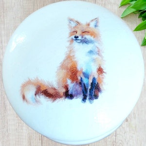 Könnte beinhalten: Eine weiße runde Keramikdose mit einem gemalten Bild eines lächelnden orangefarbenen Fuchses mit weißer Brust und schwarzen Beinen. Der Fuchs sitzt auf seinen Hinterbeinen, der Schwanz um seinen Körper gewickelt.