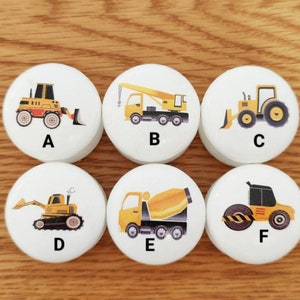 Op de afbeelding: Zes witte ronde houten tegels met daarop geprinte gele bouwvoertuigen. Elke tegel heeft een letter van A tot en met F erop gedrukt.