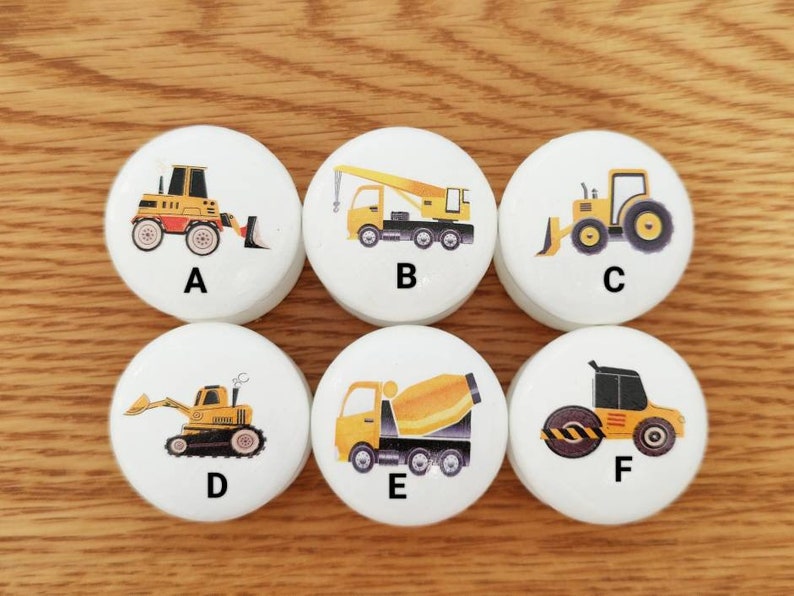 Op de afbeelding: Zes witte ronde houten tegels met daarop geprinte gele bouwvoertuigen. Elke tegel heeft een letter van A tot en met F erop gedrukt.