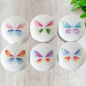 Könnte beinhalten: Sechs runde Keramikknöpfe mit einem Aquarell-Schmetterlingsdesign. Jeder Knopf hat eine andere Farbkombination aus Rosa, Blau, Gelb und Orange.
