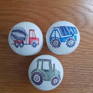 May include: Three white ceramic knobs with different construction vehicle designs. The first knob features a red and gray cement mixer truck. The second knob features a blue dump truck with gray stripes. The third knob features a green tractor.