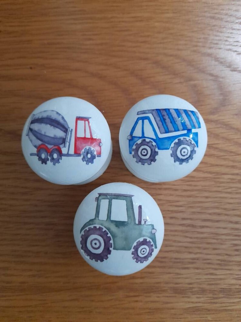 May include: Three white ceramic knobs with different construction vehicle designs. The first knob features a red and gray cement mixer truck. The second knob features a blue dump truck with gray stripes. The third knob features a green tractor.