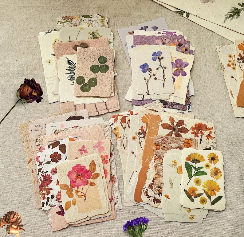 May include: A collection of handmade paper cards with pressed flowers. The cards feature various floral designs, including clovers, roses, and wildflowers. The color palette includes pinks, yellows, and greens, with a natural, rustic aesthetic. The cards are ideal for scrapbooking or crafting.