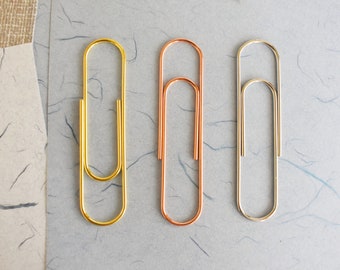 May include: Three metal paper clips in different colors: gold, copper, and silver. The paper clips are arranged in a row on a light gray background.
