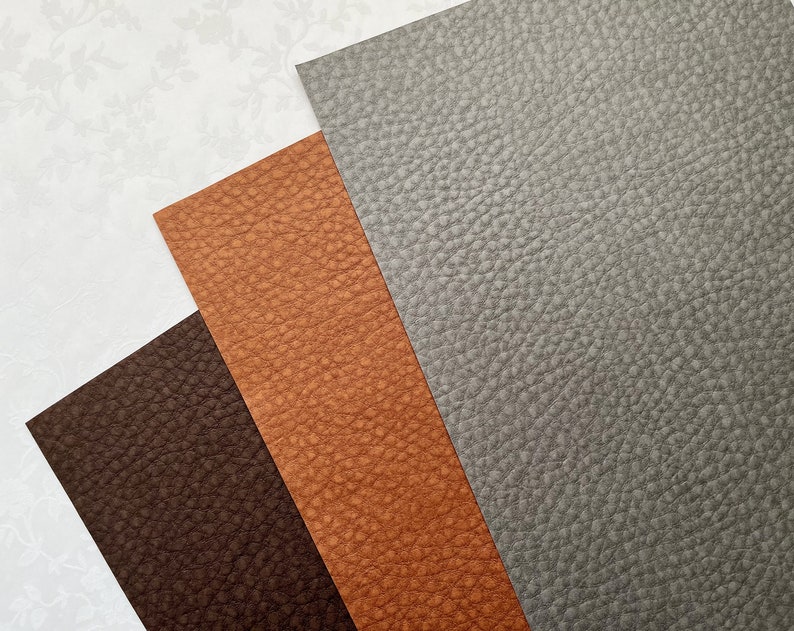 May include: Three sheets of textured faux leather in shades of brown and gray. The sheets are arranged at angles, with a dark brown sheet at the bottom, a tan sheet in the middle, and a gray sheet at the top. The background is white with a subtle floral pattern.