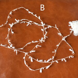 May include: A spool of white, fringed trim with a small amount of trim already unwound on a brown surface. The label on the spool reads "B".