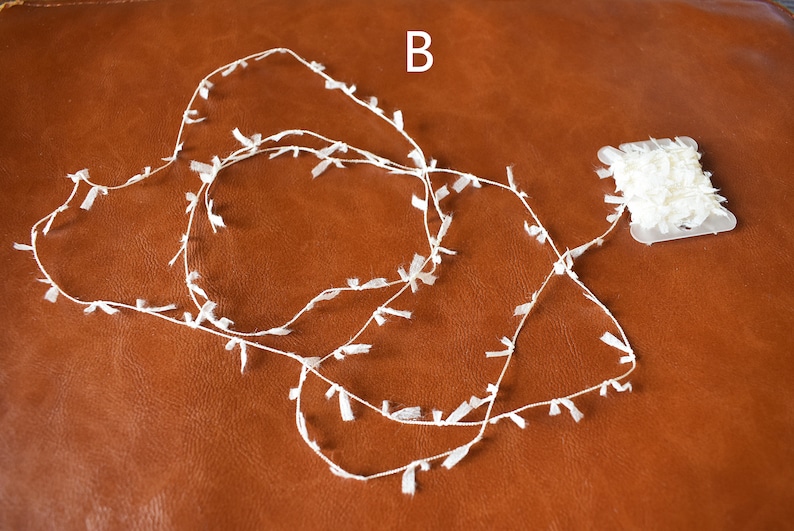 May include: A spool of white, fringed trim with a small amount of trim already unwound on a brown surface. The label on the spool reads "B".