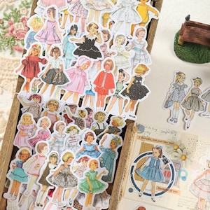 May include: A collection of vintage-style stickers featuring illustrations of girls wearing various dresses. The stickers are in a wooden box and are perfect for scrapbooking, journaling, or adding a touch of whimsy to any project.