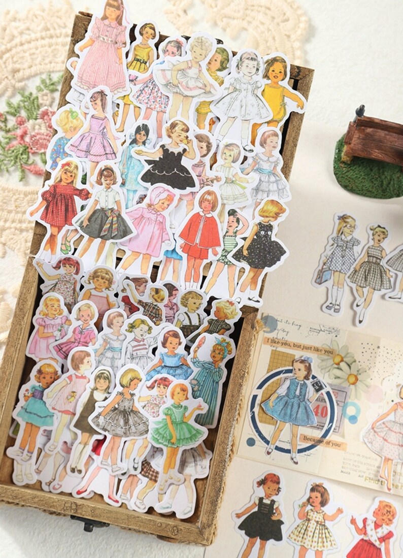 May include: A collection of vintage-style stickers featuring illustrations of girls wearing various dresses. The stickers are in a wooden box and are perfect for scrapbooking, journaling, or adding a touch of whimsy to any project.