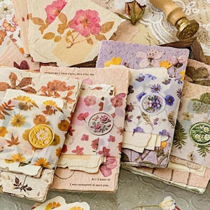 May include: A collection of handmade paper cards and envelopes, adorned with pressed flowers and botanical elements. The cards feature a variety of floral designs, including roses and daisies, with wax seals and decorative accents. The color palette includes soft pinks, creams, and natural tones.