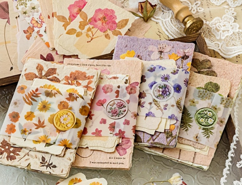 May include: A collection of handmade paper cards and envelopes, adorned with pressed flowers and botanical elements. The cards feature a variety of floral designs, including roses and daisies, with wax seals and decorative accents. The color palette includes soft pinks, creams, and natural tones.