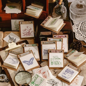 Puede incluir: Una colección de pequeños blocs de notas de estilo vintage con diversas ilustraciones de temática natural. Los blocs son de color marrón con detalles metálicos y presentan diseños como setas, mariposas y un frasco con flores. El texto en los blocs incluye "Wild Berry Tale", "Time Travel" y "Glass Garden".