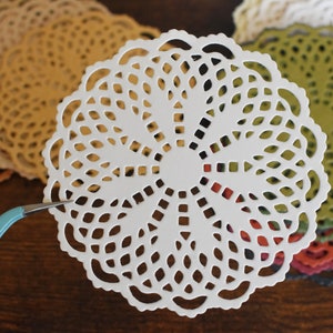 May include: A white paper doily with a delicate lace pattern. The doily is surrounded by other doilies in various colors, including brown, green, red, and black.