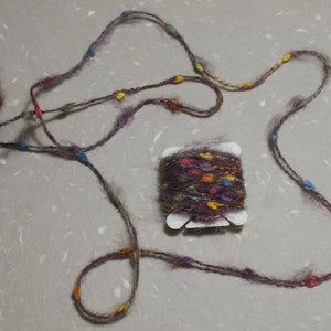 Puede incluir: Una madeja de hilo marr&oacute;n con motas multicolores, enrollada en un carrete de pl&aacute;stico blanco. El hilo tambi&eacute;n se muestra en un bucle suelto con cuentas de colores.